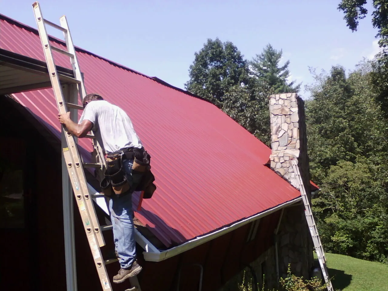 Roofer on a ladder beside a red metal roof and stone chimney — Soffit & Fascia Repair by Roofing Brothers in Chappaqua